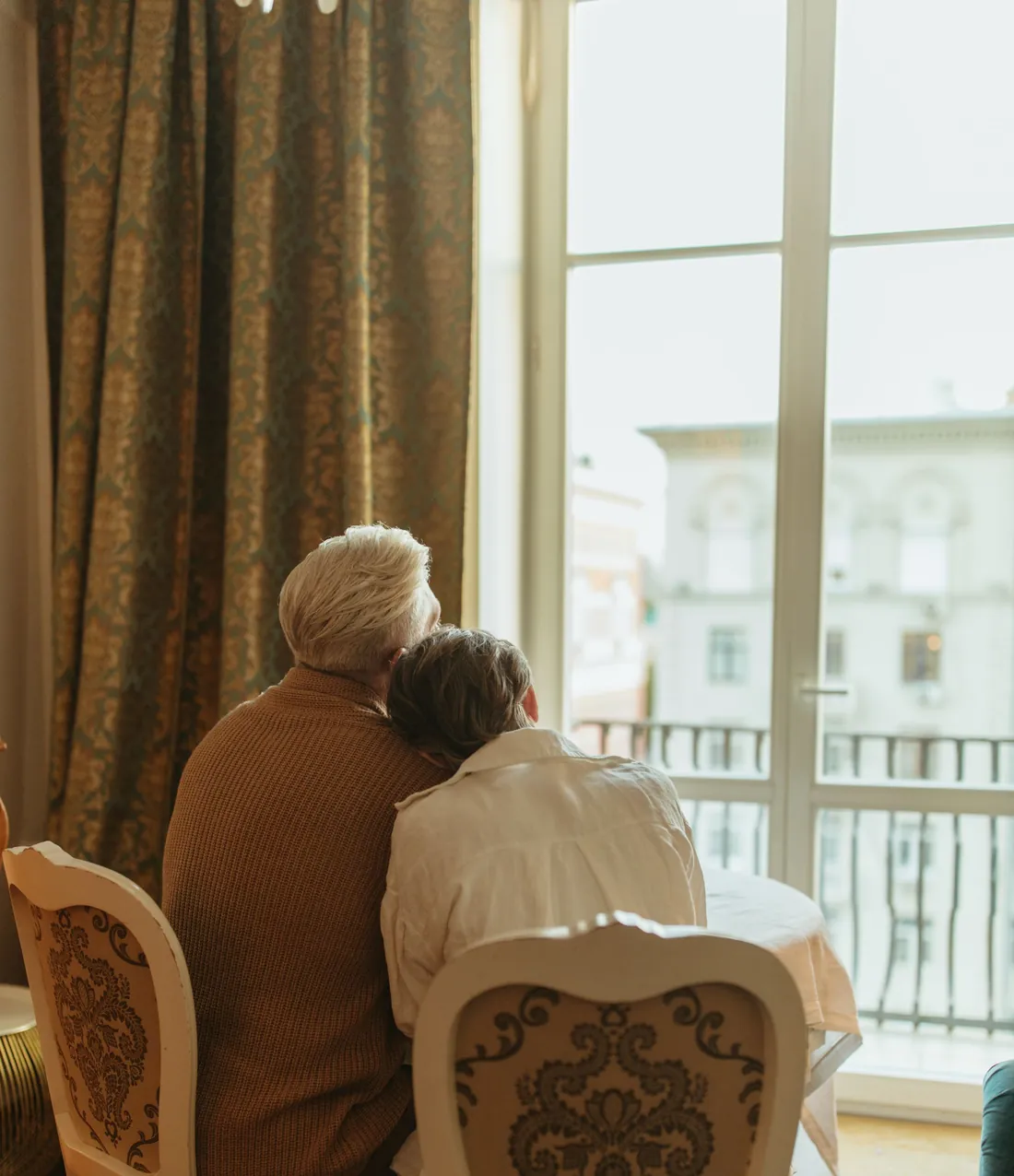 Een ouder echtpaar zit dicht tegen elkaar aan bij het raam van hun woonkamer