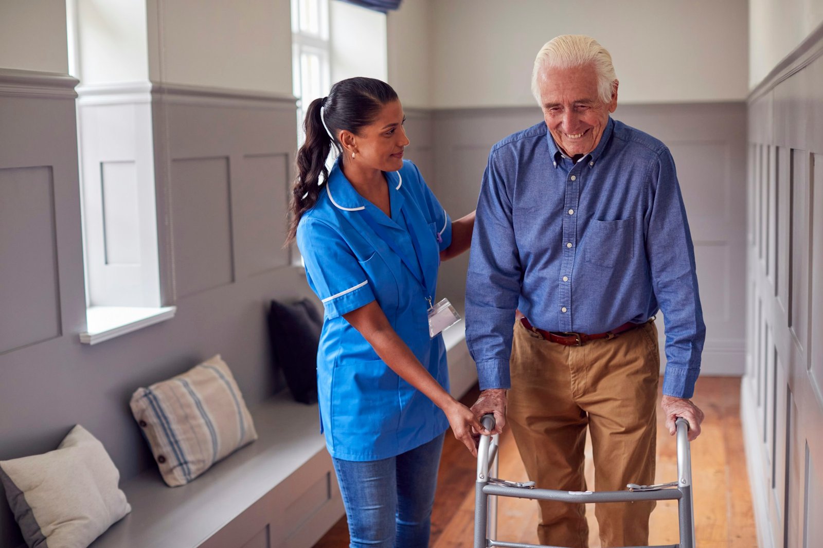 Senior Man At Home Using Walking Frame Being Helped By Female Care Worker In Uniform